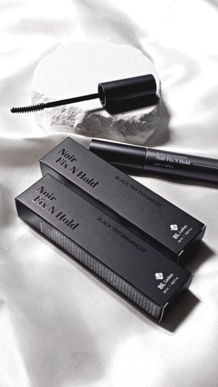 BL Lashes Noir Fix N Hold black tint enhancer with applicator brush and black product packaging arranged on white satin fabric.