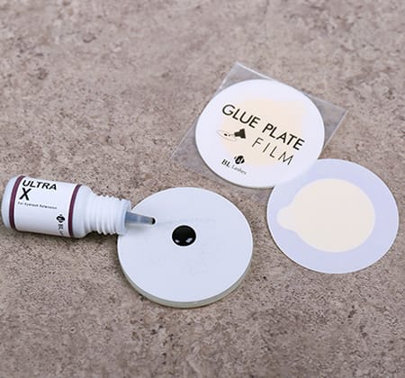 Glue plate with a drop of eyelash extension adhesive, protective glue plate films and an open adhesive bottle on a stone surface.