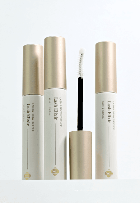 Three Lash Elixir lash and brow essence tubes with champagne-gold caps, displayed alongside an open applicator wand with a white spoolie brush on a clean white background.