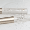 Two Lash Elixir lash and brow essence tubes with champagne-gold caps, displayed in clear gel with water droplets for a fresh, hydrating beauty aesthetic.