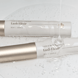 Two Lash Elixir lash and brow essence tubes with champagne-gold caps, displayed in clear gel with water droplets for a fresh, hydrating beauty aesthetic.