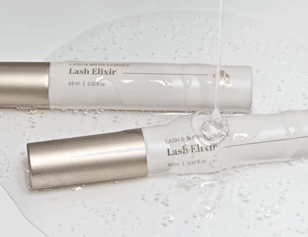 Two Lash Elixir lash and brow essence tubes with champagne-gold caps, displayed in clear gel with water droplets for a fresh, hydrating beauty aesthetic.