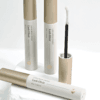 Lash Elixir lash and brow serum tubes with gold caps and an open spoolie applicator, arranged on a white stone pedestal.