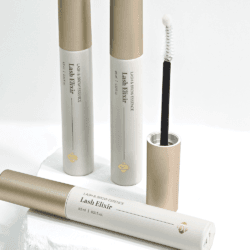 Lash Elixir Image 6 Lash Elixir lash and brow serum tubes with gold caps and an open spoolie applicator, arranged on a white stone pedestal.