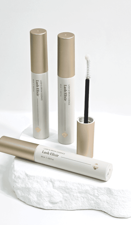 Lash Elixir lash and brow serum tubes with gold caps and an open spoolie applicator, arranged on a white stone pedestal.