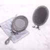 MIRROR_4 Two ornate vintage-style silver mirrors, one handheld and one freestanding, displayed on a light marble surface with a soft storage pouch.