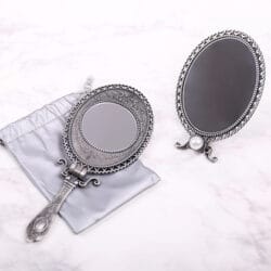 MIRROR_4 Two ornate vintage-style silver mirrors, one handheld and one freestanding, displayed on a light marble surface with a soft storage pouch.