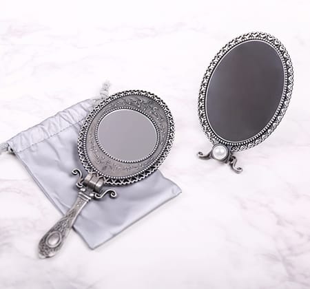 MIRROR_4 Two ornate vintage-style silver mirrors, one handheld and one freestanding, displayed on a light marble surface with a soft storage pouch.