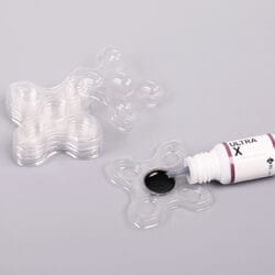 Clear plastic multi-well glue trays with black eyelash extension adhesive being dispensed from a professional lash glue bottle.