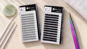 Zwei Boxen mit BL-Lashes-Wimpernextensions und eine Rainbow-Pinzette auf einem hellen Arbeitsplatz mit Jadestein.